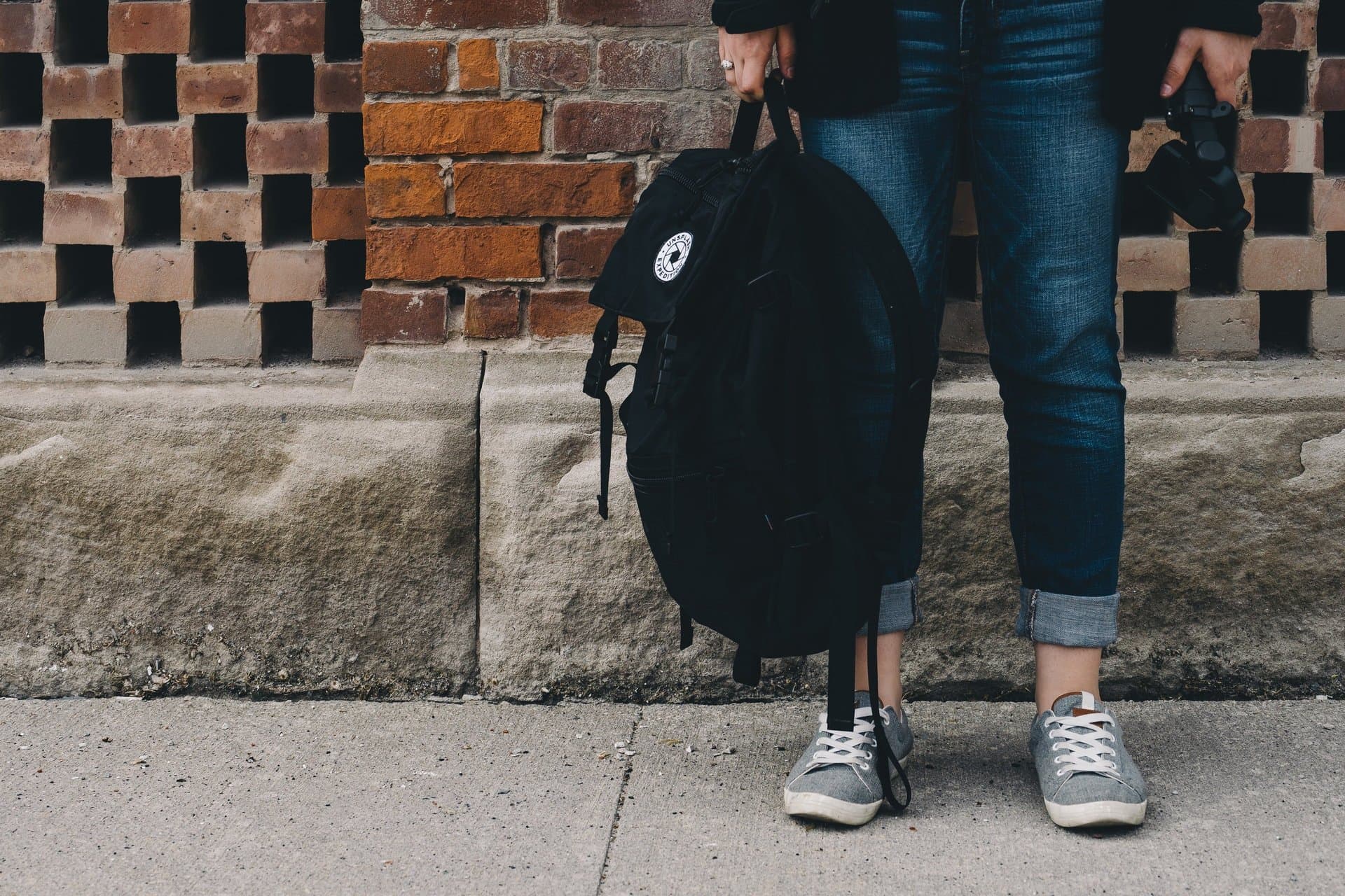 Estudiante con mochila y cámara de pie junto a una pared de ladrillo
